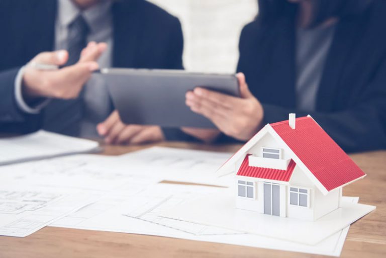 Modell eines Einfamilienhauses, im Hintergrund zeigt eine Person einer anderen Person etwas auf einem Tablet